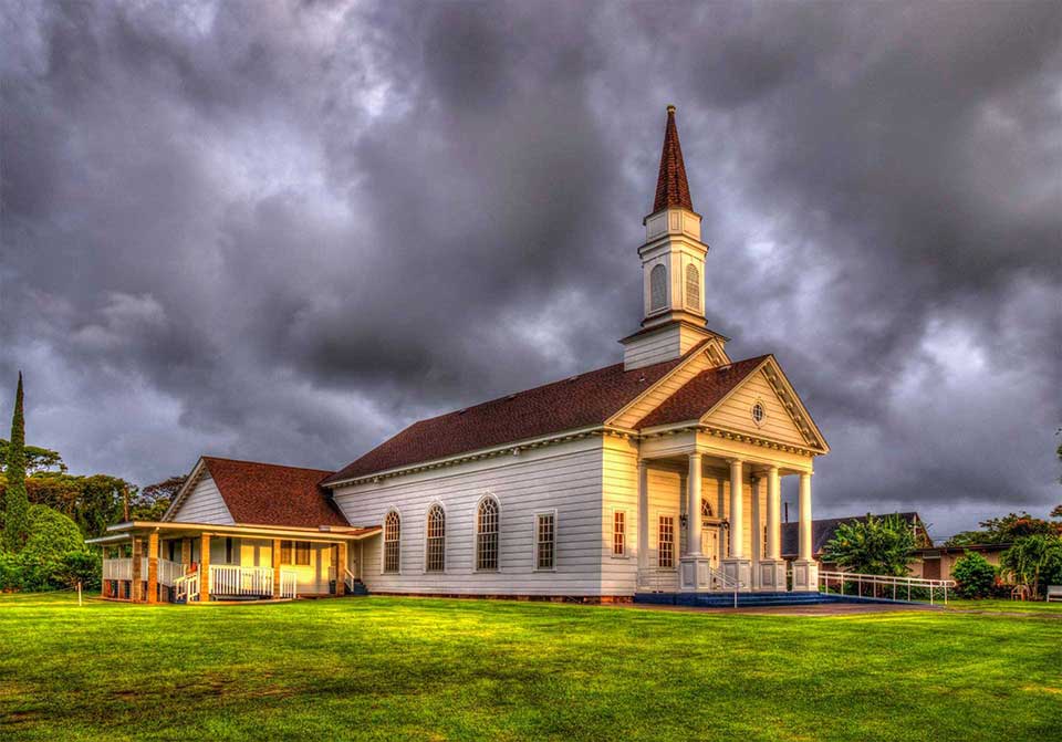 The Church at Koloa, a white historic-style church with tall columns and a steeple, illuminated beneath dramatic dark storm clouds.