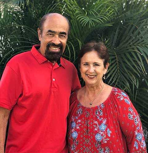 Pastors Harold and Christine Kilborn smiling together outdoors in front of lush green palm plants.
