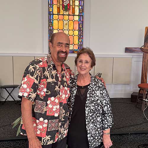 Pastors Harold and Christine Kilborn smiling together outdoors in front of the stained glass window in the pulpit.